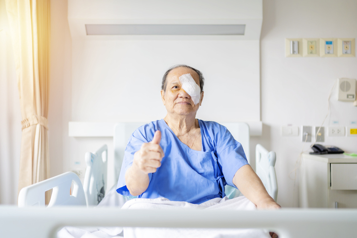 Man giving a thumbs up sitting in a hospital bed with a patch over his eye after cataract surgery