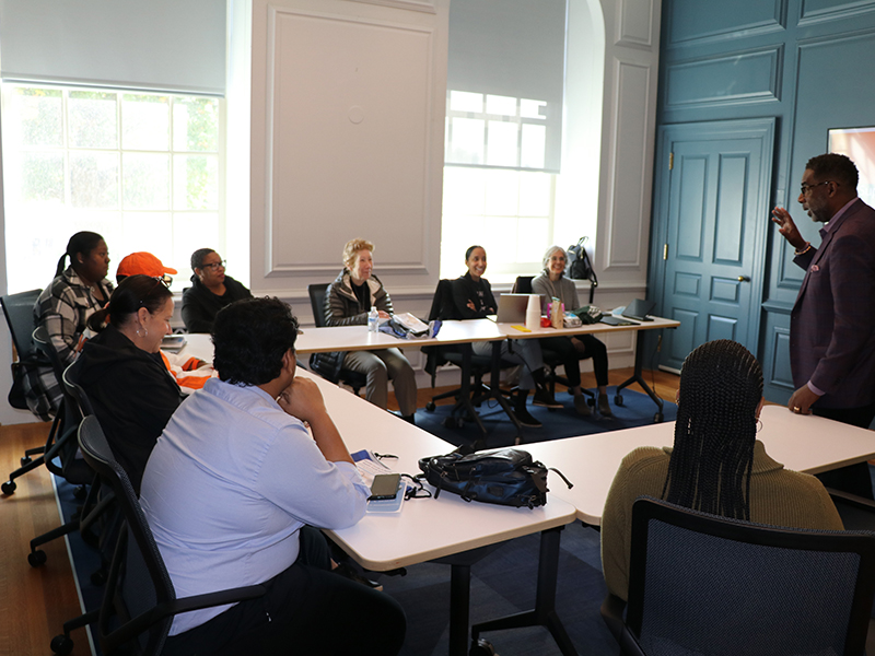 Facilitator speaks to a small group seated around tables in a bright meeting room during a workshop