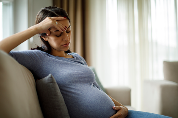 Pregnant woman sitting on a couch with eyes closed, holding her forehead and belly, appearing tired or unwell